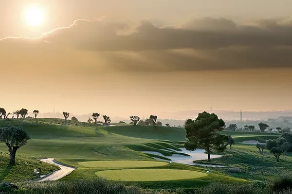 Ein malerischer Golfplatz bei Sonnenaufgang auf einer Golf-Gruppenreise nach Mallorca, mit sanften, grünen Fairways, Sandbunkern, vereinzelten Bäumen und einem dunstigen Himmel, der von der aufgehenden Sonne beleuchtet wird, die teilweise von Wolken verdeckt ist.