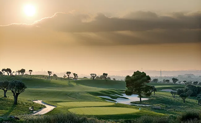 Ein malerischer Golfplatz bei Sonnenaufgang auf einer Golf-Gruppenreise nach Mallorca, mit sanften, grünen Fairways, Sandbunkern, vereinzelten Bäumen und einem dunstigen Himmel, der von der aufgehenden Sonne beleuchtet wird, die teilweise von Wolken verdeckt ist.