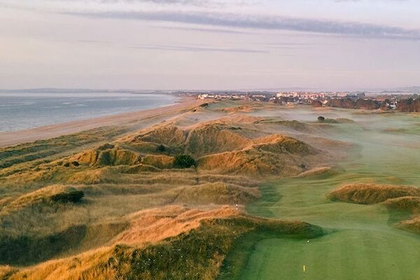 Ein malerischer Blick auf einen Golfplatz an der Küste mit sanften Dünen, grünen Fairways und Nebelschwaden, die im frühen Morgenlicht aufsteigen – die perfekte Inspiration für Ihre Packliste für einen Golfurlaub in Irland. Unter einem leicht bewölkten Himmel sind die Küste und Gebäude in der Ferne zu sehen.