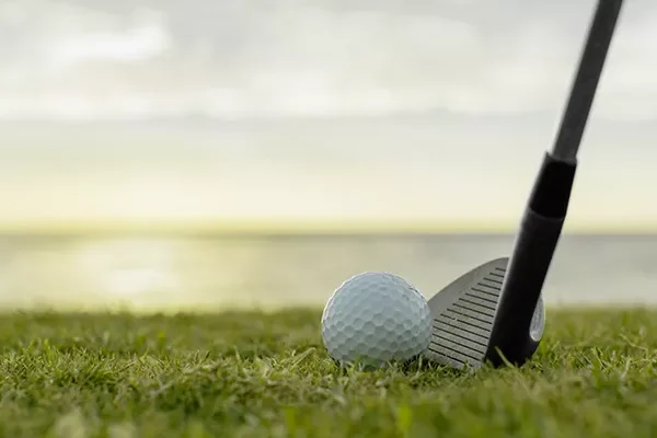 Eine Nahaufnahme eines Golfschlägers, der neben einem Golfball auf dem Rasen liegt, mit einem unscharfen Hintergrund aus Wasser und Himmel bei Sonnenuntergang – perfekt für einen Erfahrungsbericht über eine Golfreise nach Ägypten.