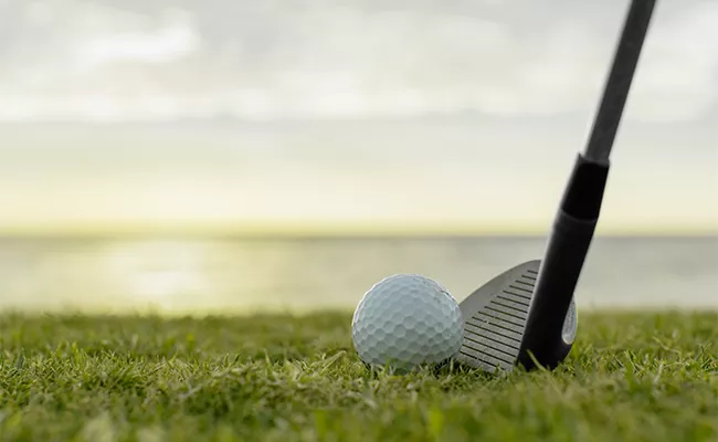 Eine Nahaufnahme eines Golfschlägers, der neben einem Golfball auf dem Rasen liegt, mit einem unscharfen Hintergrund aus Wasser und Himmel bei Sonnenuntergang – perfekt für einen Erfahrungsbericht über eine Golfreise nach Ägypten.