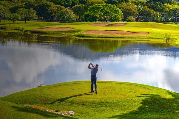 packliste-costa-rica Ein Golfer schwingt seinen Schläger auf einem üppig grünen Platz in der Nähe eines ruhigen Sees, im Hintergrund sind Sandbunker und Bäume zu sehen, während der Himmel teilweise bewölkt ist.