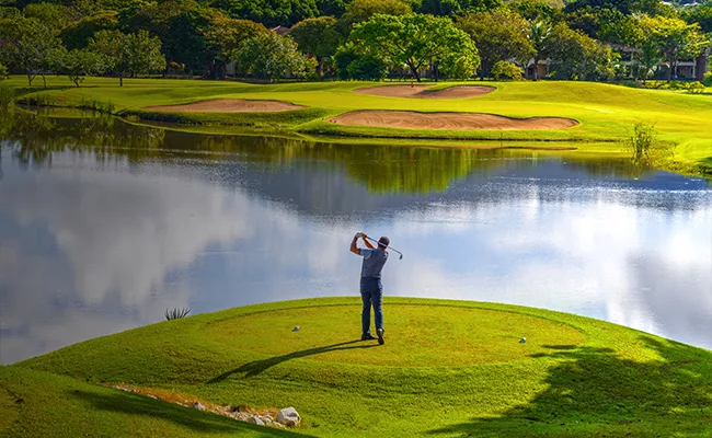 packliste-costa-rica Ein Golfer schwingt seinen Schläger auf einem üppig grünen Platz in der Nähe eines ruhigen Sees, im Hintergrund sind Sandbunker und Bäume zu sehen, während der Himmel teilweise bewölkt ist.