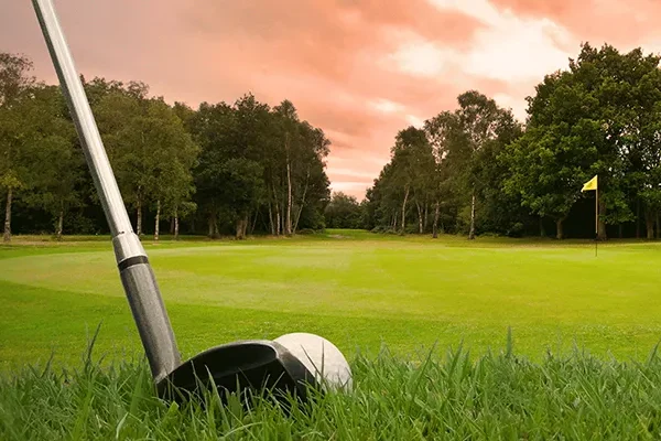 Ein Golfschläger liegt auf dem Rasen in der Nähe eines Grüns, eine gelbe Flagge markiert das Loch, und Bäume säumen den Platz unter einem dramatischen, bewölkten Himmel bei Sonnenuntergang – ein Bild, das die landschaftliche Schönheit der Golfplätze in Irland widerspiegelt.