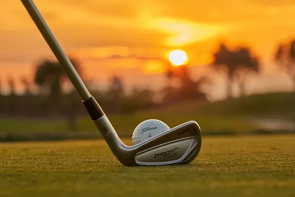 Ein Golfschläger, der bereit ist, einen Titleist-Golfball während einer Golf-Gruppenreise auf Mallorca auf das Grün zu schlagen, vor dem Hintergrund eines leuchtend orangefarbenen Sonnenuntergangs und verschwommener Bäume.