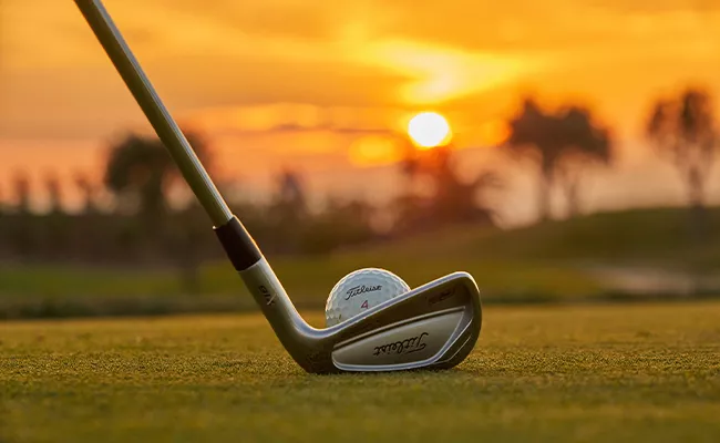 Ein Golfschläger, der bereit ist, einen Titleist-Golfball während einer Golf-Gruppenreise auf Mallorca auf das Grün zu schlagen, vor dem Hintergrund eines leuchtend orangefarbenen Sonnenuntergangs und verschwommener Bäume.