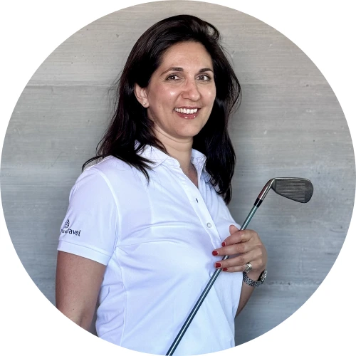 Anne Franz, mit langen dunklen Haaren und weißem Poloshirt, hält einen Golfschläger in der Hand und lächelt in die Kamera, während sie vor einer grauen Wand steht.