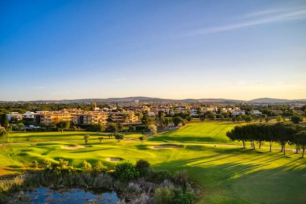 Ein malerischer Ausblick, der an einen Reisebericht über eine Golfreise nach Portugal erinnert: gepflegte Grüns, Sandbunker, Bäume und nahegelegene Wohnhäuser unter strahlend blauem Himmel bei Sonnenuntergang.