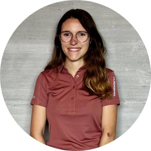 Lara Kellenberger, eine Frau mit langen braunen Haaren und Brille, die ein rosafarbenes Hemd mit Kragen trägt, steht vor einer grauen Betonwand und lächelt in die Kamera.