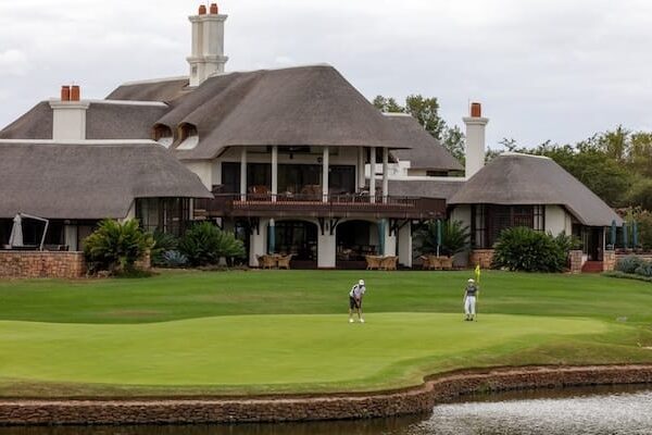 Leopard Creek Zwei Menschen spielen Golf auf einem Grün in der Nähe eines Teiches vor einem großen Clubhaus mit Strohdach, umgeben von viel Grün und bewölktem Himmel – eine idyllische Szene, die an Reiseziele erinnert, die man auf einer gut vorbereiteten Packliste für Südafrika finden könnte.