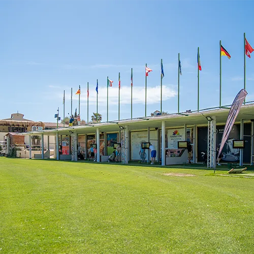 Eine Reihe von überdachten und bevölkerten Golfplätzen bietet die perfekte Kulisse für Individuelle Trainingsreisen, über denen mehrere internationale Flaggen wehen, und eine weite grüne Wiese unter einem klaren blauen Himmel.