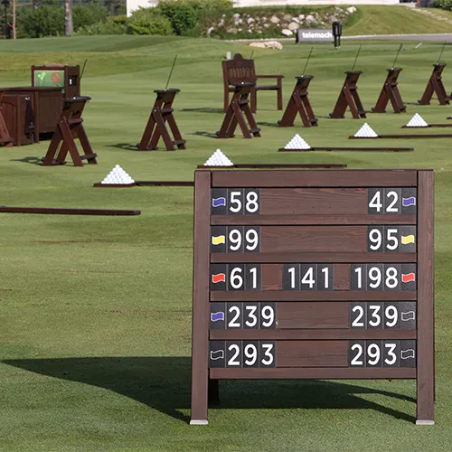 Ein hölzerner Entfernungsmesser zeigt die Entfernungen vor mehreren Übungsstationen an, an denen Golfbälle in Pyramiden auf grünem Gras gestapelt sind - perfekt für Golfer, die individuelle Trainingsreisen planen.