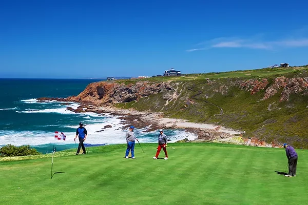 Vier Menschen spielen unter einem klaren, sonnigen Himmel auf einem Grün in der Nähe einer Küstenklippe Golf, mit blauen Meereswellen und einer zerklüfteten Küstenlinie im Hintergrund – genau wie die malerischen Golfplätze, für die Südafrika bekannt ist.