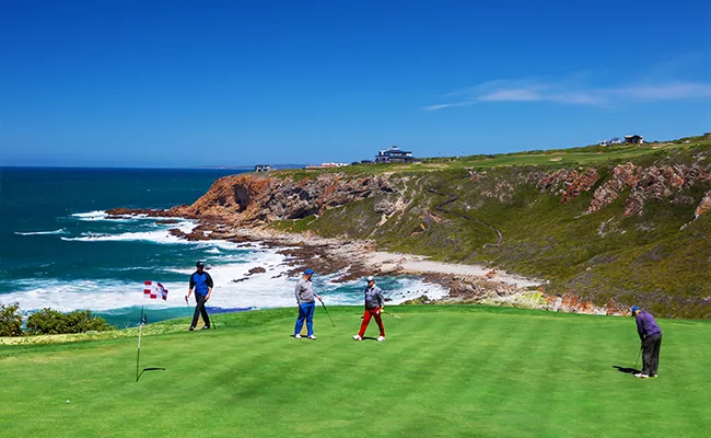 golf-anfaengerreise-suedafrika-2 Vier Menschen spielen unter einem klaren, sonnigen Himmel auf einem Grün in der Nähe einer Küstenklippe Golf, mit blauen Meereswellen und einer zerklüfteten Küstenlinie im Hintergrund – genau wie die malerischen Golfplätze, für die Südafrika bekannt ist.