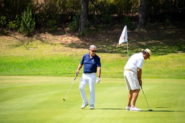 An einem sonnigen Tag spielen zwei Männer Golf; der eine, in weißer Kleidung und mit weißem Hut, puttet, während der andere, der ein dunkelblaues Hemd und eine weiße Hose trägt, daneben zusieht. Auf dem Grün weht eine Golffahne – eine ideale Szene auf den berühmten Golfplätzen Portugals.