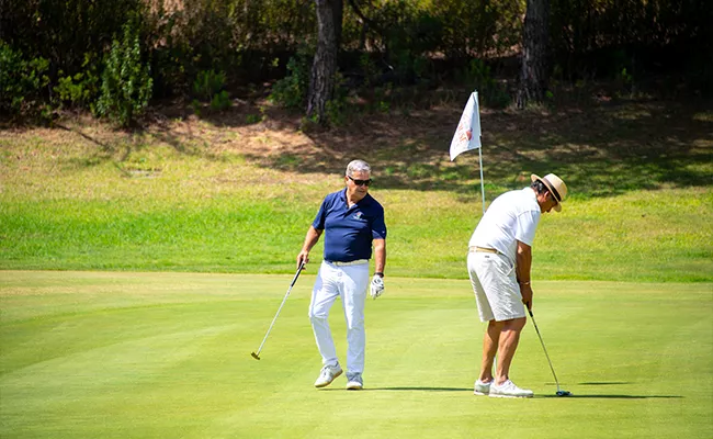 golf-gruppenreise-portugal-2 An einem sonnigen Tag spielen zwei Männer Golf; der eine, in weißer Kleidung und mit weißem Hut, puttet, während der andere, der ein dunkelblaues Hemd und eine weiße Hose trägt, daneben zusieht. Auf dem Grün weht eine Golffahne – eine ideale Szene auf den berühmten Golfplätzen Portugals.