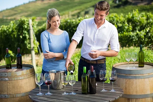 Ein Mann schenkt Wein in ein Glas ein, während eine Frau ihm dabei zusieht; beide stehen an Weinfässern und einem Tisch mit Flaschen und Gläsern in einem Weinberg an einem sonnigen Tag – ein Erlebnis, das ebenso unvergesslich ist wie die Erkundung der Golfplätze in Südafrika.