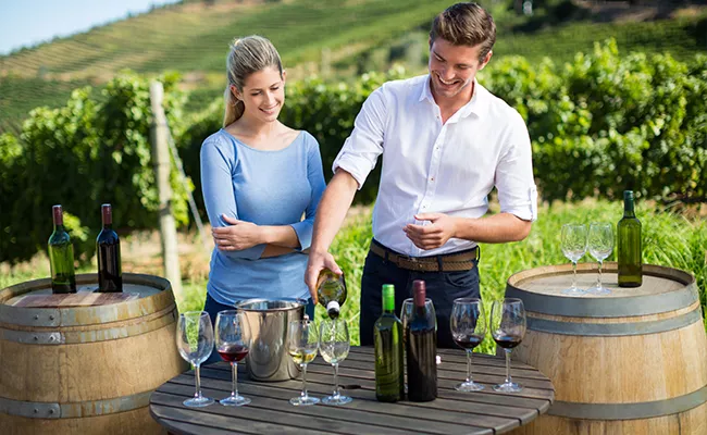 golf-gruppenreise-suedafrika-2 Ein Mann schenkt Wein in ein Glas ein, während eine Frau ihm dabei zusieht; beide stehen an Weinfässern und einem Tisch mit Flaschen und Gläsern in einem Weinberg an einem sonnigen Tag – ein Erlebnis, das ebenso unvergesslich ist wie die Erkundung der Golfplätze in Südafrika.