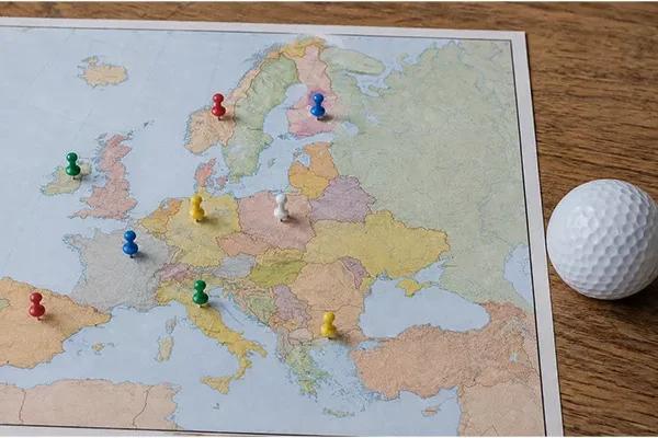 golfplaetze-europa Eine Europakarte mit bunten Stecknadeln, die verschiedene Orte markieren, liegt auf einer Holzfläche neben einem weißen Golfball auf der rechten Seite.