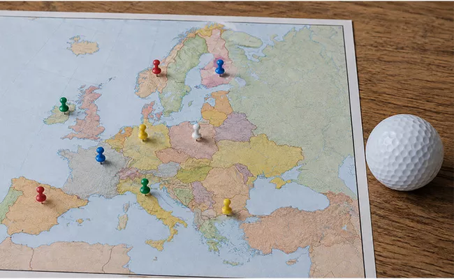 golfplaetze-europa Eine Europakarte mit bunten Stecknadeln, die verschiedene Orte markieren, liegt auf einer Holzfläche neben einem weißen Golfball auf der rechten Seite.