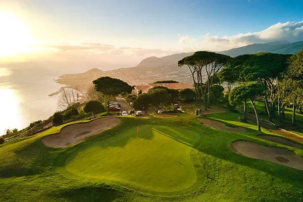 Ein malerischer Golfplatz liegt an einem üppig grünen Hang mit Blick auf den Ozean bei Sonnenuntergang und erinnert an die atemberaubenden Golfplätze, die Portugal zu bieten hat – mit Bäumen, Sandbunkern und fernen Bergen unter einem teilweise bewölkten Himmel.
