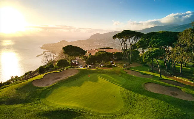 golfplaetze-portugal Ein malerischer Golfplatz liegt an einem üppig grünen Hang mit Blick auf den Ozean bei Sonnenuntergang und erinnert an die atemberaubenden Golfplätze, die Portugal zu bieten hat – mit Bäumen, Sandbunkern und fernen Bergen unter einem teilweise bewölkten Himmel.