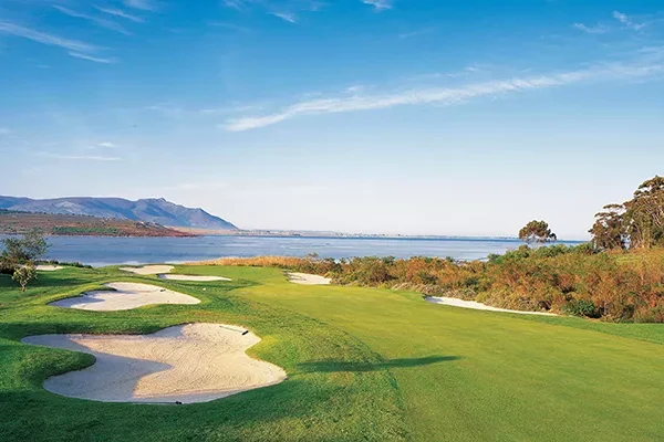 Ein malerischer Golfplatz, der an die atemberaubenden Golfplätze Südafrikas erinnert, mit Sandbunkern, üppig grünen Fairways und einem Blick auf einen ruhigen See oder das Meer, eingerahmt von Bergen und Bäumen unter einem strahlend blauen Himmel.