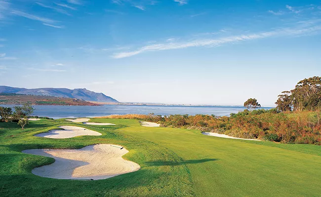 golfplaetze-suedafrika Ein malerischer Golfplatz, der an die atemberaubenden Golfplätze Südafrikas erinnert, mit Sandbunkern, üppig grünen Fairways und einem Blick auf einen ruhigen See oder das Meer, eingerahmt von Bergen und Bäumen unter einem strahlend blauen Himmel.