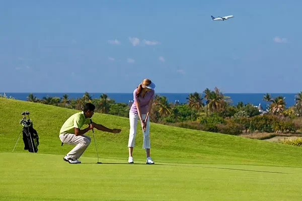 golfreise-inkl-greenfee-und-flug Ein Mann und eine Frau spielen Golf auf einem grünen, von Palmen gesäumten Platz, mit Blick auf das Meer und ein fernes Flugzeug - ideal für eine Golfreise inkl. Greenfee und Flug unter einem klaren blauen Himmel.