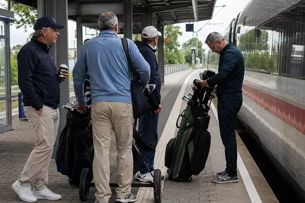 nachhaltige-golfreise-planen Vier Männer mit Golftaschen stehen auf einem Bahnsteig in der Nähe eines stehenden Zuges. Ein Mann überprüft seine Golftasche, während die anderen in Freizeitkleidung und mit Hut dastehen und sich unterhalten. Im Hintergrund sind Bäume und Bahngleise zu sehen.