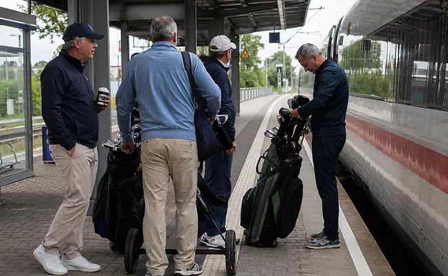 Vier Männer mit Golftaschen stehen auf einem Bahnsteig in der Nähe eines stehenden Zuges. Ein Mann überprüft seine Golftasche, während die anderen in Freizeitkleidung und mit Hut dastehen und sich unterhalten. Im Hintergrund sind Bäume und Bahngleise zu sehen.