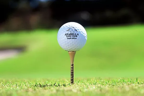 Eine Nahaufnahme eines Golfballs auf einem Tee, beide mit der Aufschrift „Arabella“, auf dem leuchtend grünen Rasen eines der renommierten Golfplätze Südafrikas, mit sanft unscharfem Hintergrund.