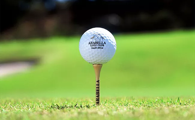 Eine Nahaufnahme eines Golfballs auf einem Tee, beide mit der Aufschrift „Arabella“, auf dem leuchtend grünen Rasen eines der renommierten Golfplätze Südafrikas, mit sanft unscharfem Hintergrund.