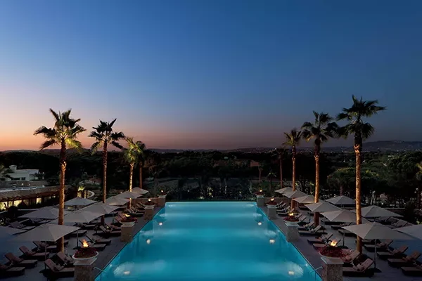Ein Infinity-Pool, umgeben von Liegestühlen und Palmen bei Sonnenuntergang, mit Blick auf malerische Golfplätze in Portugal vor dem Hintergrund eines sich verdunkelnden Himmels.