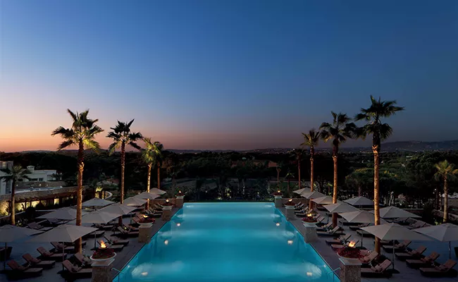 reisebericht-portugal-2 Ein Infinity-Pool, umgeben von Liegestühlen und Palmen bei Sonnenuntergang, mit Blick auf malerische Golfplätze in Portugal vor dem Hintergrund eines sich verdunkelnden Himmels.
