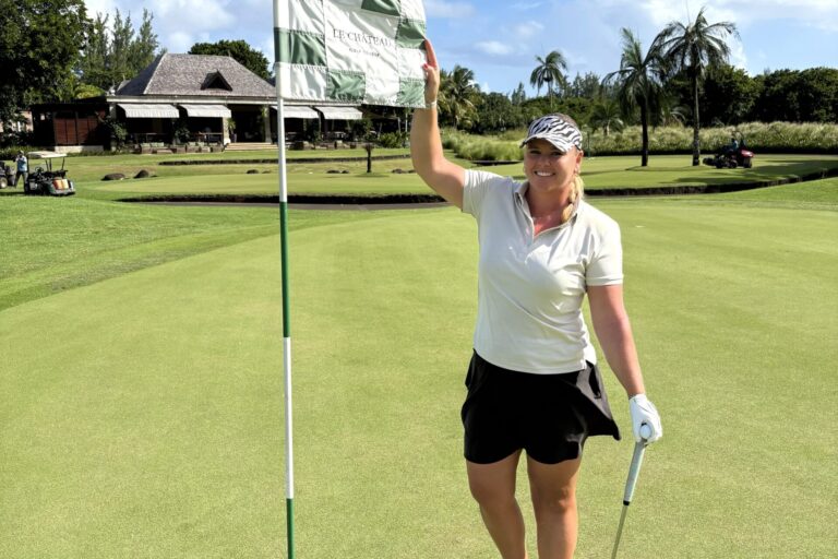 Eine Frau in Golfkleidung steht lächelnd auf einem Grün neben einem Flaggenstock und hält einen Golfschläger in der Hand. Im Hintergrund sind unter blauem Himmel ein Clubhaus, Bäume und Palmen zu sehen.