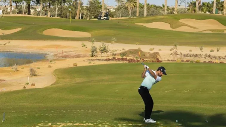 Ein Golfer in hellblauem Hemd und schwarzer Hose schwingt seinen Schläger auf einem grünen Golfplatz und vermittelt dabei den malerischen Charme einer Golfrundreise durch Irland – mit Sanddünen, Palmen, einem kleinen Teich und einem Golfwagen im Hintergrund.