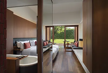 Ein modernes Hotelzimmer mit Holzböden, einem großen Bett, einer Sitzecke vor raumhohen Fenstern und Blick auf einen grünen Garten draußen. Im Vordergrund ist teilweise eine Badewanne zu sehen.