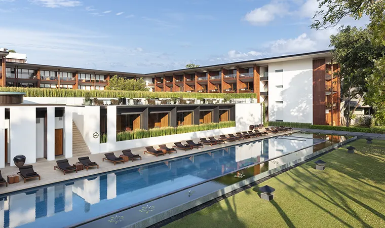 Modernes Resort mit einem langen, rechteckigen Swimmingpool, umgeben von Liegestühlen und viel Grün. Das mehrstöckige Gebäude verfügt über Balkone und große Fenster und hebt sich vor einem blauen Himmel mit vereinzelten Wolken ab.