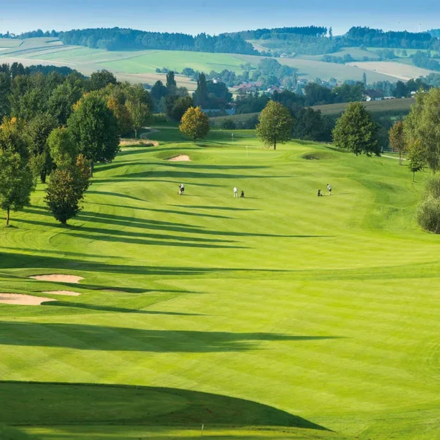 Bad-Griesbach-Golf Ein malerischer Golfplatz in Kernen-Schönried mit sanft geschwungenen, grünen Fairways, Sandbunkern, vereinzelten Bäumen und Golfern in der Ferne, die unter klarem Himmel spielen, umgeben von üppiger Landschaft und bewaldeten Hügeln.
