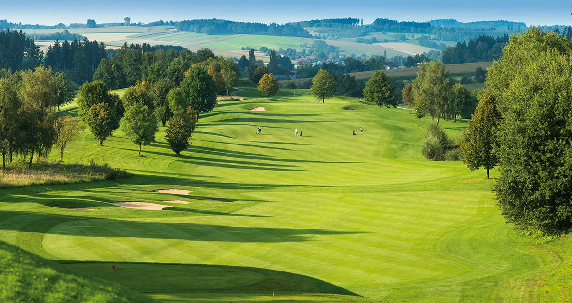 Ein malerischer Golfplatz in Kernen-Schönried mit sanft geschwungenen, grünen Fairways, Sandbunkern, vereinzelten Bäumen und Golfern in der Ferne, die unter klarem Himmel spielen, umgeben von üppiger Landschaft und bewaldeten Hügeln.