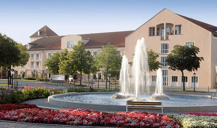 Bad-Griesbach-Hotel-760x450-1 Vor dem großen, hellen Gebäude von Kernen Schönried mit seinem rotgedeckten Dach steht unter strahlend blauem Himmel ein runder Springbrunnen mit Wasserfontänen, umgeben von bunten Blumenbeeten und Bäumen.