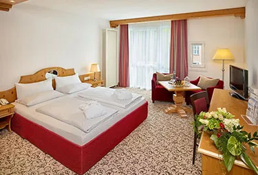 Bad-Griesbach-Hotel-Zimmer-375x255-1 Ein helles Hotelzimmer im „Kernen Schönried“ verfügt über ein Doppelbett mit weißer Bettwäsche, einen Schreibtisch mit Blumen, einen Fernseher, einen kleinen Esstisch mit Stühlen, einen Sessel, große Fenster mit Vorhängen und einen gemusterten Teppich.