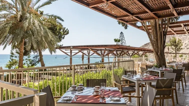 Eine Restaurantterrasse im Freien mit gedeckten Tischen und Blick auf das Meer – der perfekte Ort, um sich nach einer Golfrundreise durch Irland zu entspannen, umgeben von Palmen und viel Grün, mit einer hölzernen Pergola, die Schatten spendet und einen malerischen Blick auf die Küste bietet.
