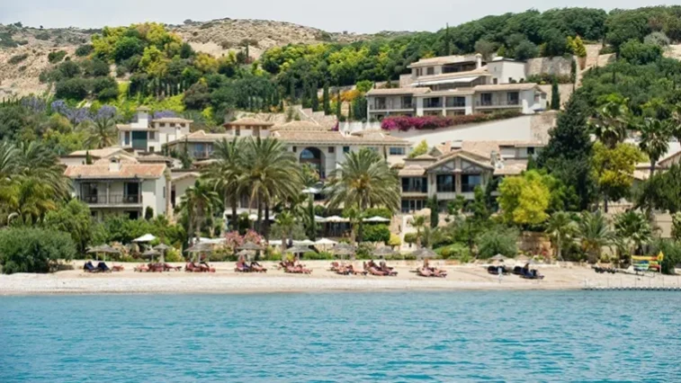 Ein Luxusresort mit Villen, umgeben von üppigem Grün und Palmen, direkt an einem Sandstrand mit Liegestühlen und Sonnenschirmen – ideal zum Entspannen nach einer abenteuerlichen Golfrundreise durch Irland, an einem sonnigen Tag vom Wasser aus gesehen.