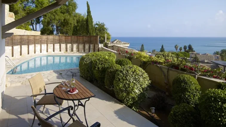 Eine Terrasse mit einem kleinen Tisch und Stühlen bietet einen Blick auf einen Swimmingpool, gepflegte Sträucher und eine malerische Aussicht auf das Meer – ideal zum Entspannen nach einer Golfrundreise durch Irland –, vor dem Hintergrund von Küstenhäusern und einem strahlend blauen Himmel.