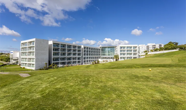 CrownPlaza-Caparica-exterior-760x450 Ein großes, modernes, weißes Hotel mit vielen Balkonen steht auf einer üppig grünen Wiese unter strahlend blauem Himmel – ein einladender Zwischenstopp auf Ihrer Irland-Rundreise entlang der Ostküste.