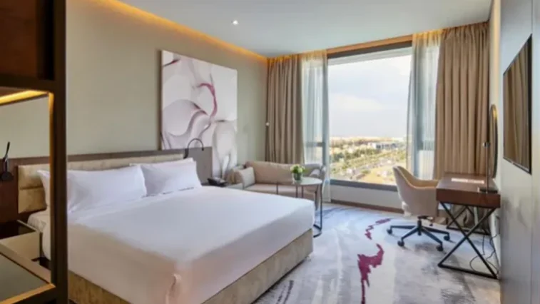 Ein modernes Hotelzimmer mit einem großen Bett, abstrakten Wandbildern, einem Sofa, einem Schreibtisch, einem Stuhl, einem Fernseher und einem großen Fenster mit Blick auf die Stadt – ideal zum Entspannen nach Ihrer Golfrundreise durch Irland. Neutrale Farbtöne und natürliches Licht schaffen eine klare, einladende Atmosphäre.