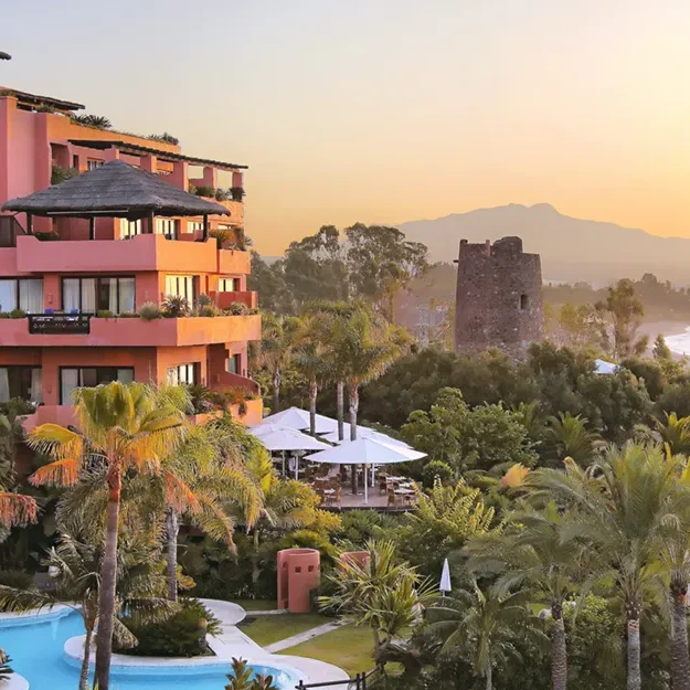 Das Melia Hacienda del Conde, ein Resort in leuchtendem Rosa mit üppigen Palmen und einem gewundenen Pool, bietet bei Sonnenuntergang einen Blick auf die malerische Küste, wobei im Hintergrund die Berge und das Meer zu sehen sind.