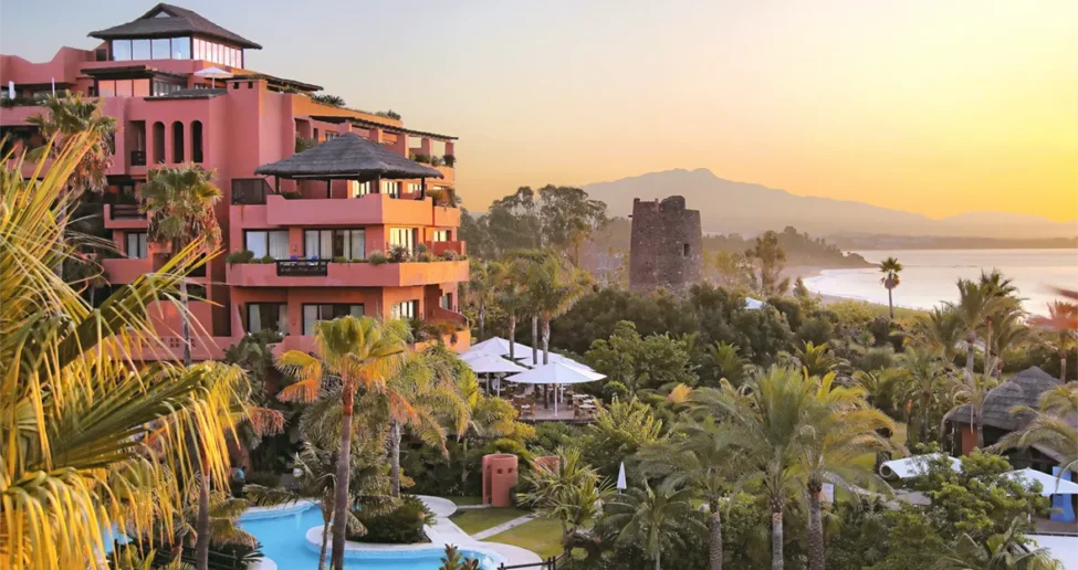 Das Melia Hacienda del Conde, ein Resort in leuchtendem Rosa mit üppigen Palmen und einem gewundenen Pool, bietet bei Sonnenuntergang einen Blick auf die malerische Küste, wobei im Hintergrund die Berge und das Meer zu sehen sind.
